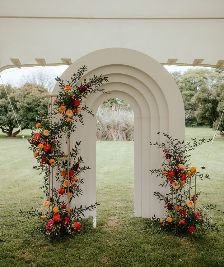 A white arched ceremony backdrop dressed with coral, orange and pink flowers on a lawn
