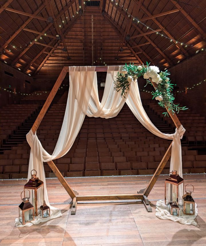 A hexagonal wooden ceremony arch dressed with cream drape, white flowers and copper lanterns